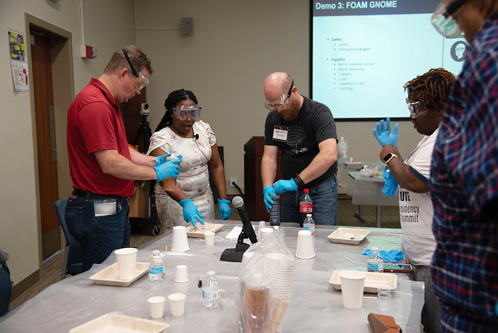 Workshop participants stand around a table donning safety gear like glasses and gloves as they prepare to practice an experiment.