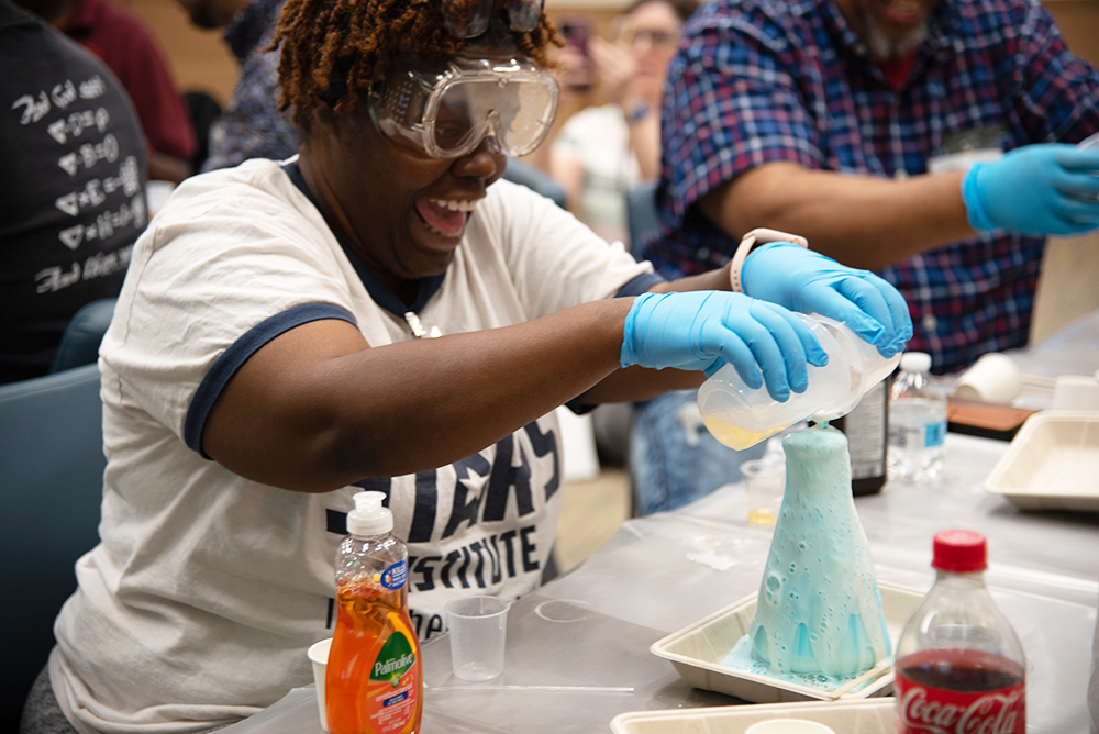 Kandice Taylor, in safety glasses, laughs as she pours liquids into a glass flask that is overflowing with foam.