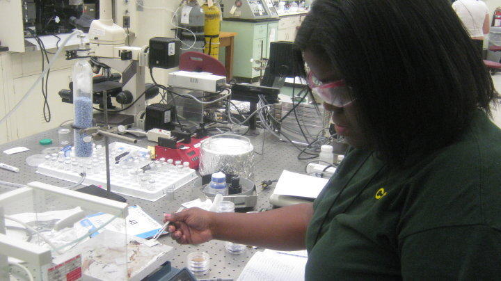 A woman in safety glasses uses tweezers to manipulate small items on a lab table. 