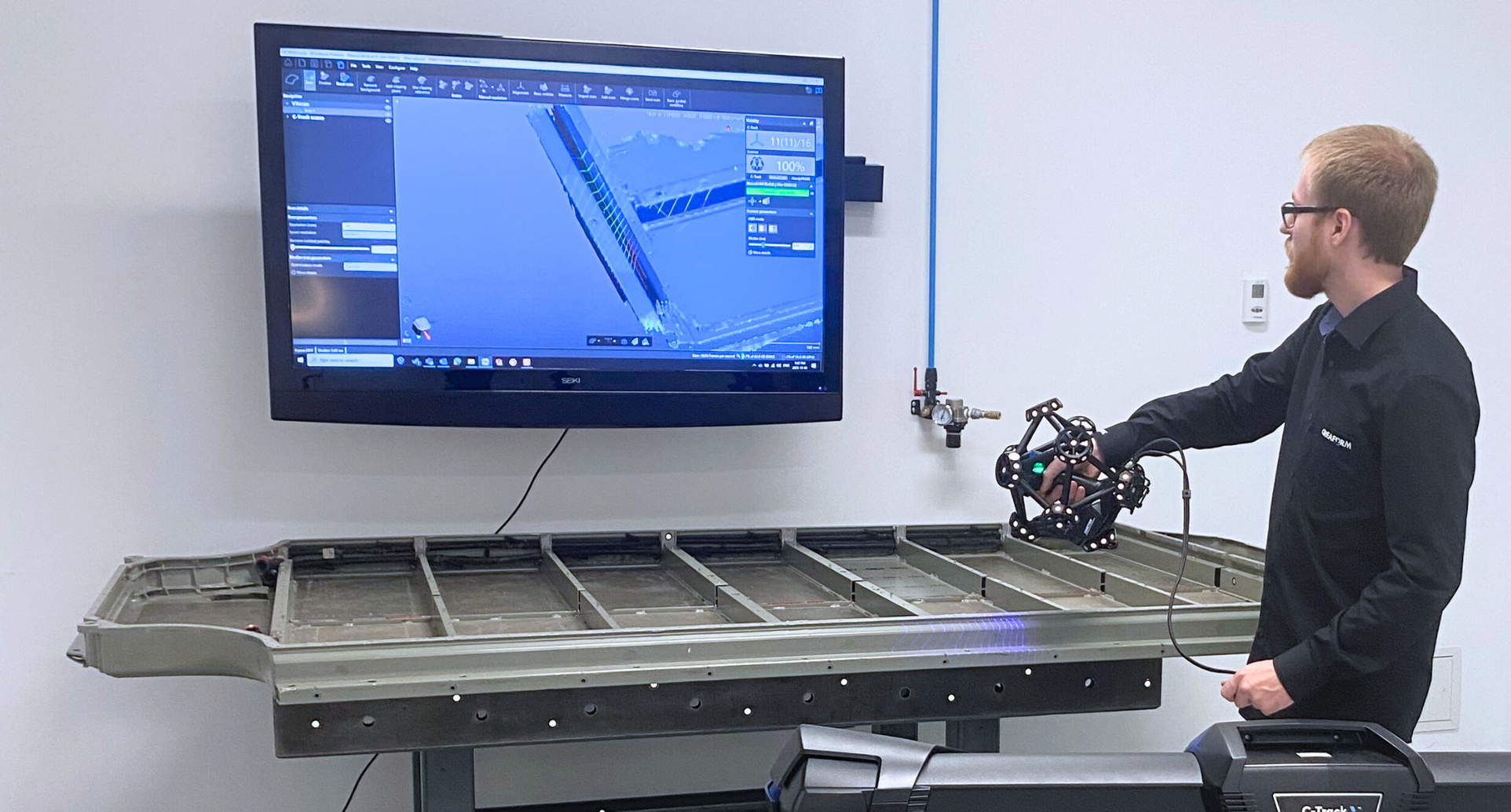 Man wearing a Creaform black shirt uses the MetraSCAN to measure an EV battery tray with C-Track in the foreground while the scan is simultaneously showing on a screen in the VXelements software.